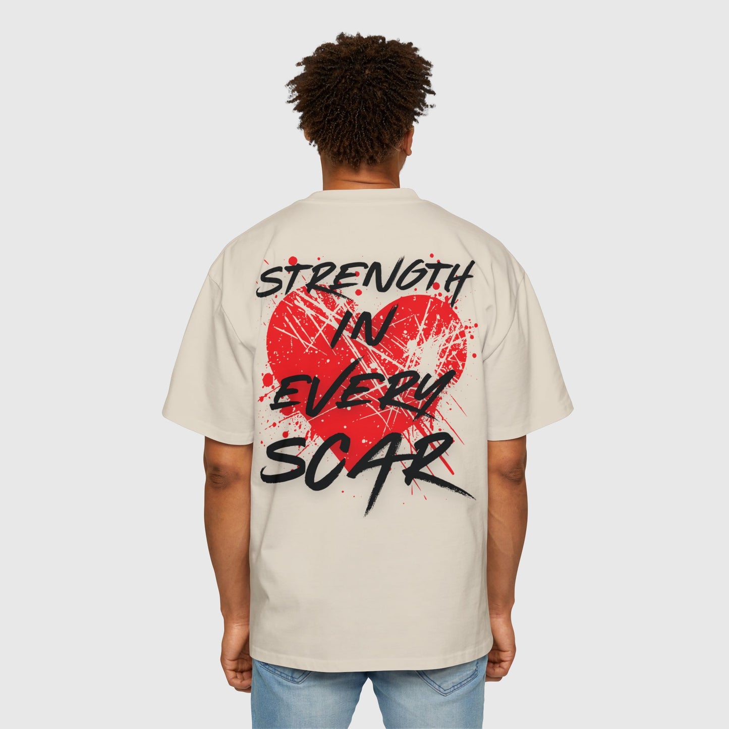 A man in a beige oversized T-shirt featuring a red splattered heart and the motivational slogan 'Strength in Every Scar' on the back. This striking design reflects active street wear that motivates and inspires confidence through bold, artistic statements