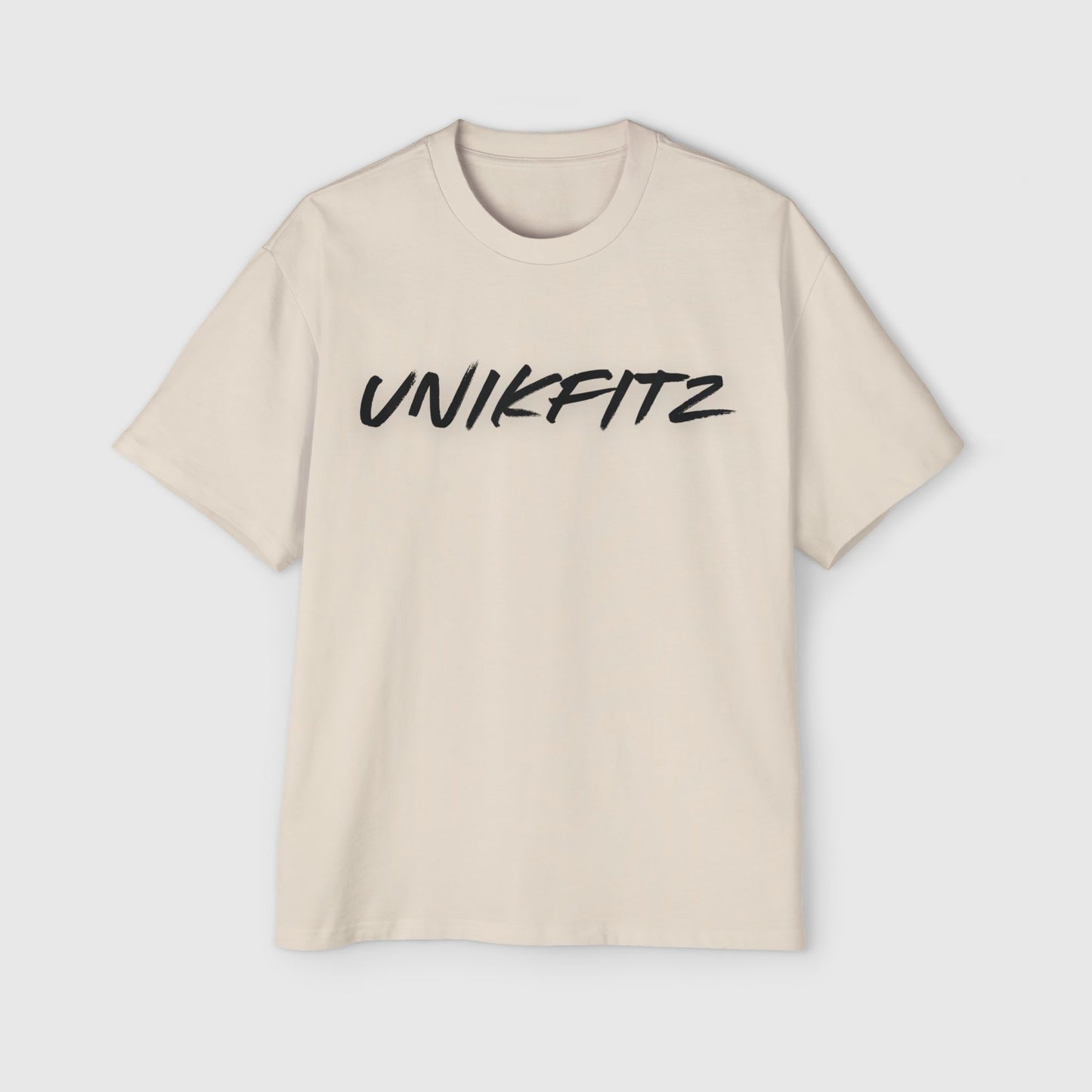 A beige oversized T-shirt displayed flat with 'UNIKFITZ' printed across the chest in bold black brushstroke font. This active street wear for the fearless misfits combines simplicity and bold design, ideal for those with a daring, confident style.