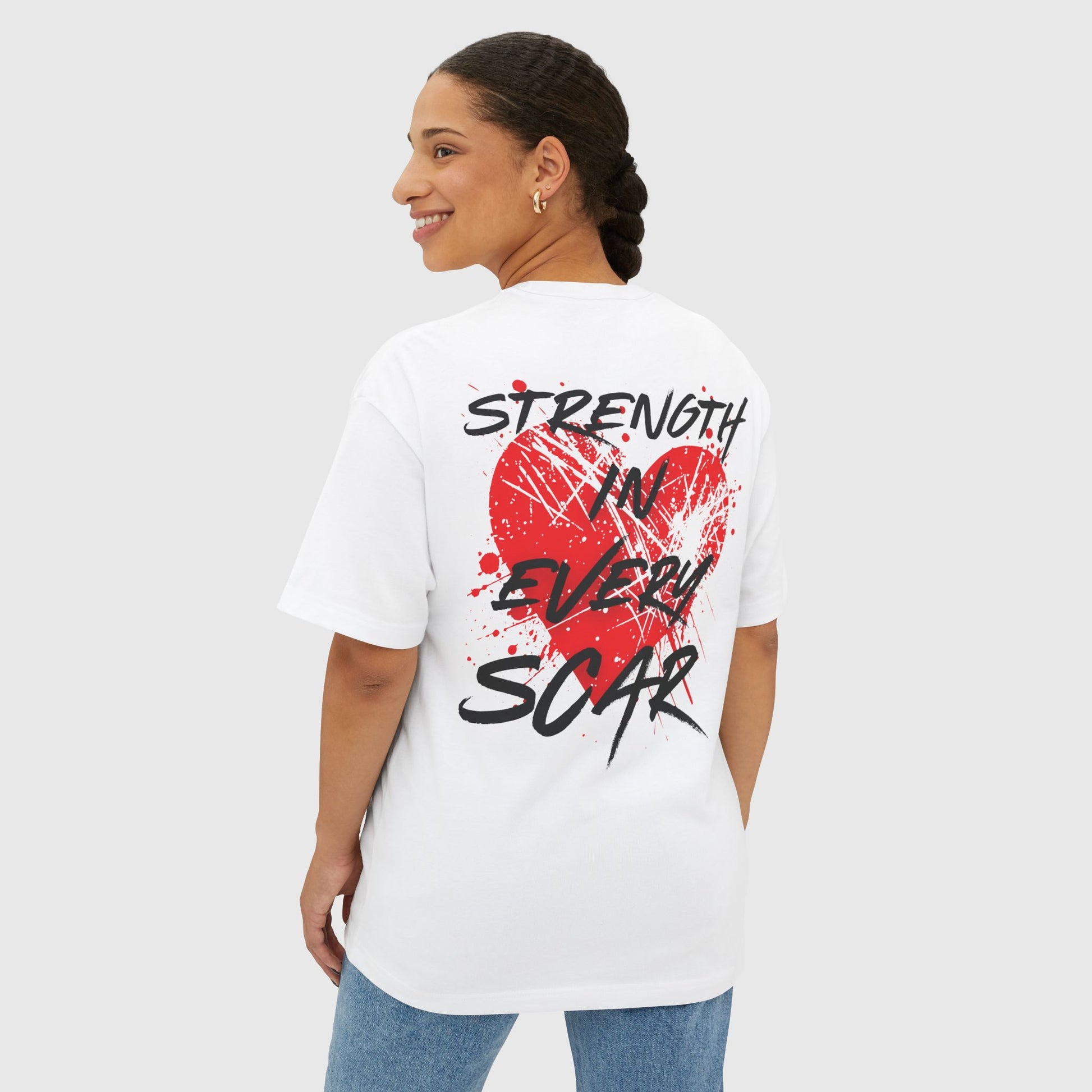 A woman wearing a white oversized T-shirt with a bold red heart and splatter design on the back, featuring the motivational slogan 'Strength in Every Scar' in black, edgy font. This active street wear that motivates combines comfort and empowerment for modern, confident individuals.