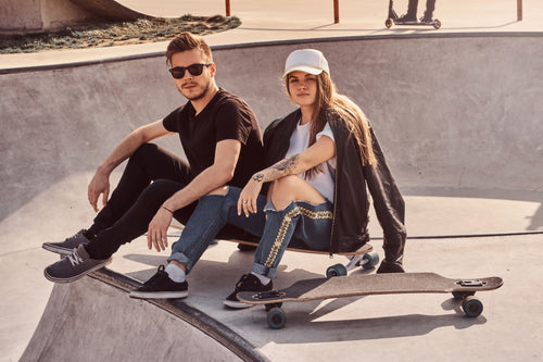 Two stylish young adults sitting at a skatepark wearing trendy casual outfits. The man sports a sleek black t-shirt and sunglasses, embodying modern men's clothing style. The woman, wearing a white t-shirt under a black leather jacket, combines it with ripped jeans and a baseball cap for a chic streetwear look. Their laid-back vibe highlights hoodie essentials and contemporary men's and women's clothing trends, perfect for casual outings