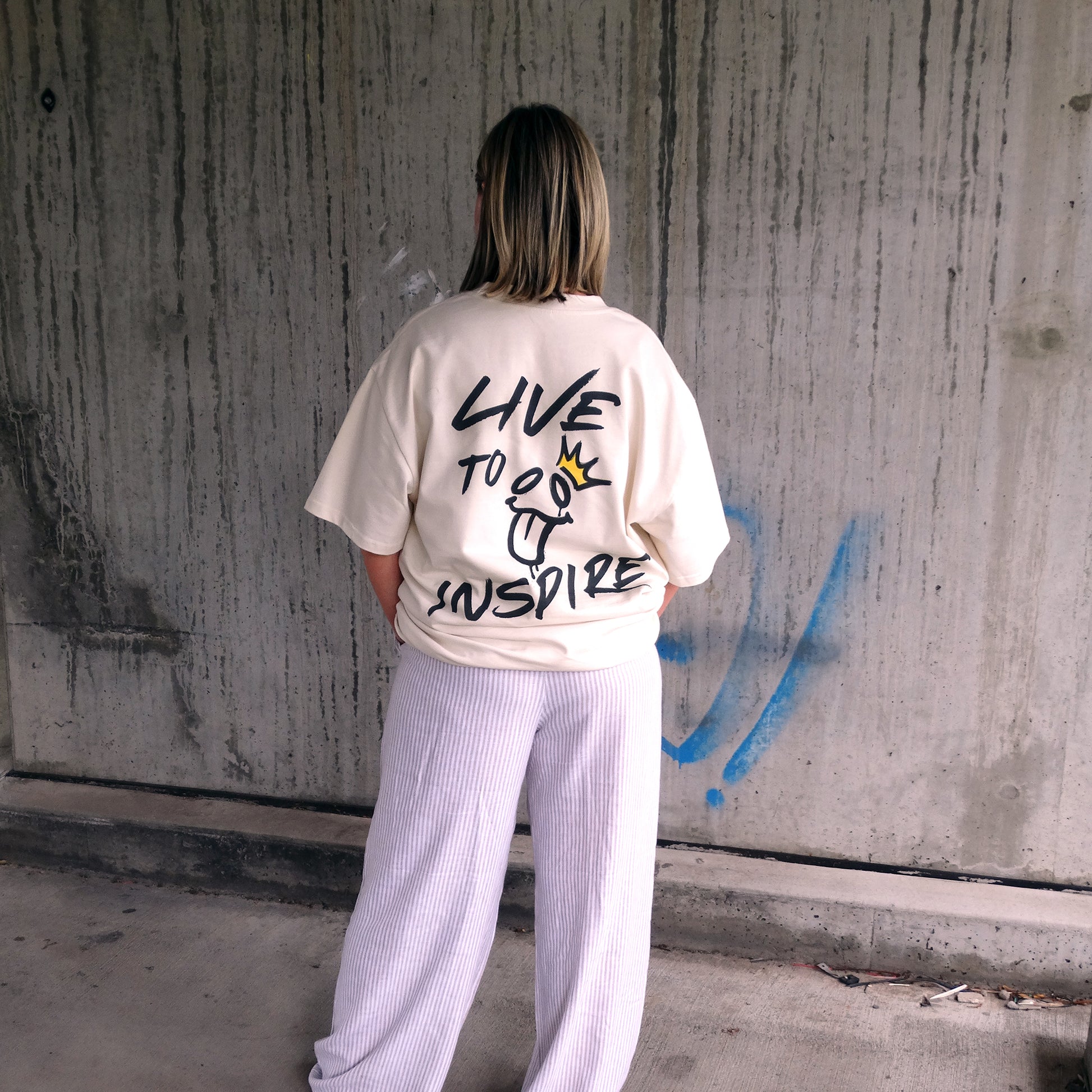 Back view of the 'Live to Inspire' women's t-shirt by UNIKFITZ, showcasing bold graffiti-style typography and a playful face graphic with a gold crown. This women's t-shirt essential combines Australian streetwear culture with a powerful message, making it a must-have for those seeking unique women's wear.